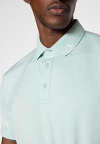 Lyseblå polo skjorte med korte ermer, knappestolpe og krage. Har en liten hvit logo på kragen og et teksturert mønster på ermene.