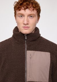 Brown fleece jacket featuring a high collar, front zipper, and a lighter brown chest pocket with a horizontal zipper. Textured fabric.