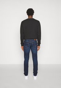 Man som står med ryggen mot kameran, iklädd en svart sweatshirt, åtsittande mörkblå jeans och vita sneakers mot en helt vit bakgrund.