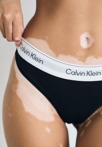 Crne badiće s bijelim Calvin Klein logotipom na struku, izrađene od glatkog materijala i s niskim strukom. Koža pokazuje različite tonove.