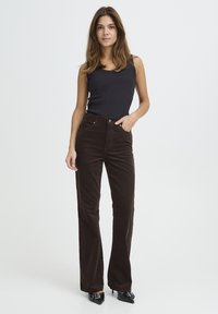 Brown corduroy flared pants with front pockets, high waist, and textured fabric, paired with a fitted black tank top and pointed black heels.