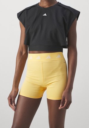 Personne portant un haut court noir sans manches et un short de sport jaune taille haute, tous deux avec des logos Adidas blancs.