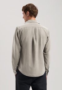 Cămașă lungă cu mânecă, de culoare gri deschis, realizată dintr-un material moale. Are un manșon cu nasturi, pliu la spate și un hem rotunjit. Design simplu și curat.