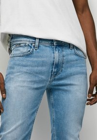 Ljust blå denimjeans med slim fit, med femficksdesign, subtila whiskering-effekter och en svart logotagg vid midjan.