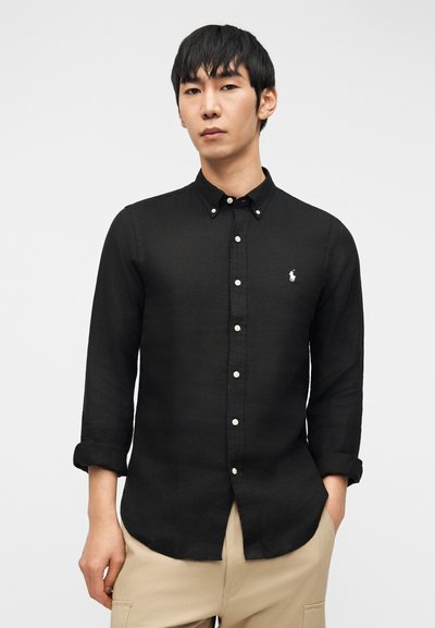 Bărbat purtând o cămașă neagră cu mâneci lungi nasturate și cu manșetele suflecate și pantaloni bej, stând în fața unui fundal simplu, deschis la culoare.