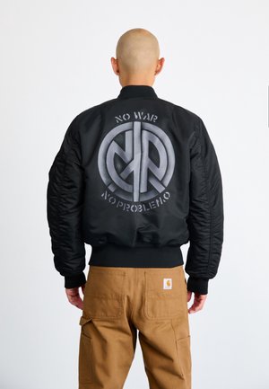 Person mit rasiertem Kopf, trägt eine schwarze Bomberjacke mit dem Aufdruck „NO WAR NO PROBLEMO“ und einem Friedenssymbol auf dem Rücken, kombiniert mit braunen Hosen.
