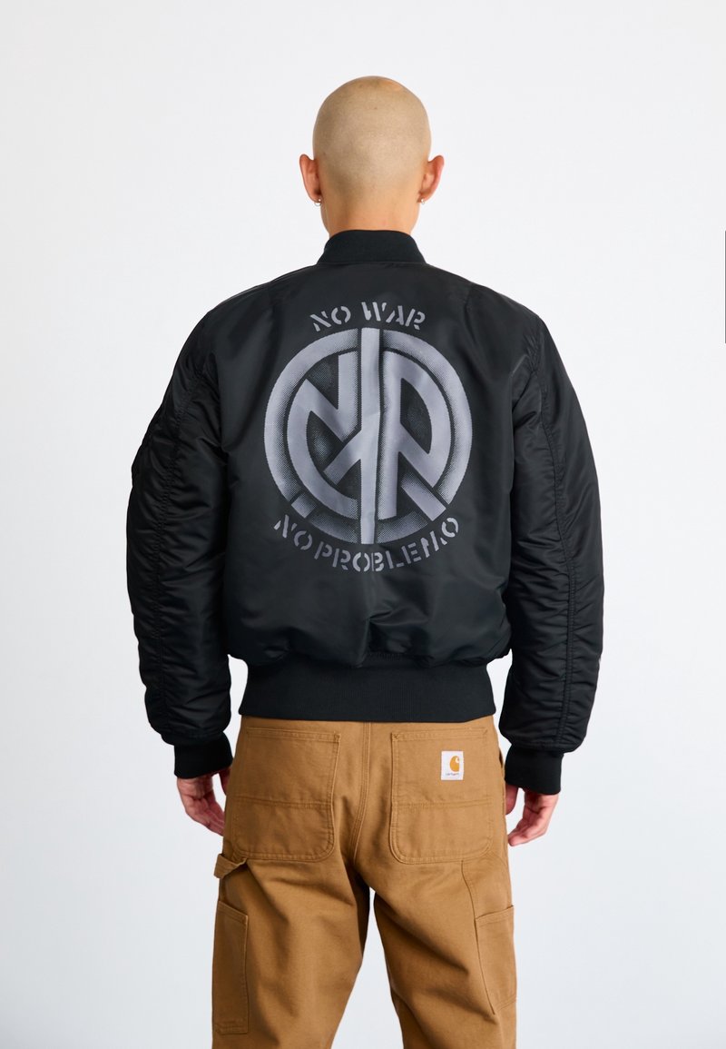 Personne à la tête rasée portant une veste bomber noire avec le texte « NO WAR NO PROBLEMO » et un symbole de paix dans le dos, associée à un pantalon marron.
