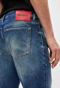 Lys blå denimjeans med en slitt finish. Har en rød skinnmerkelapp merket "HUGO" og standard baklommer.