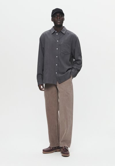 Graues Hemd mit Knöpfen und Tasche, beige weite Hose und dunkle Schuhe. Zubehör umfasst eine schwarze Mütze. Einfarbig, minimale Muster.