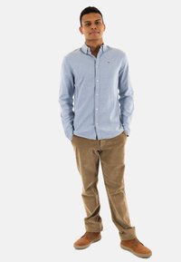 Camisa de botón azul claro, pantalones de pana marrón y zapatos marrón. La camisa tiene un cuello y un discreto detalle de logo; corte clásico.
