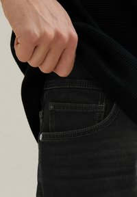 Czarne dżinsy o gładkiej fakturze, z przednią kieszenią i detalami w postaci przeszyć, obok teksturowany czarny sweter, częściowo je zakrywający.