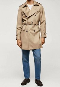  kaki trenchcoat met dubbelknopen, brede revers, ceintuur om de taille en schouderepauletten, gecombineerd met blauwe jeans en bruine schoenen.