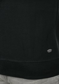 Czarny sweter o fakturze, z prążkowanym wykończeniem u dołu, z małą metalową metką z logo z boku. Tkanina ma skośny wzór pasków.