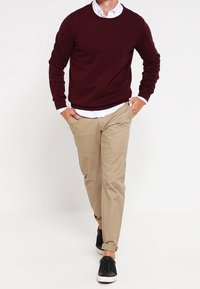 Bordeaux gebreide trui over een witte overhemd, gecombineerd met tan chino's en zwarte instapschoenen. Heeft opgerolde mouwen en een minimaal logo.