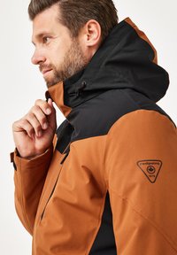 Braune und schwarze wasserdichte Jacke mit hohem Kragen und verstellbarem Kapuzen. Verfügt über Reißverschlusstaschen und einen Logo-Patch am Ärmel.