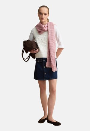 Joven mujer con camisa blanca, chaleco bordado, pañuelo rosa, falda vaquera oscura, bailarinas negras, sosteniendo un bolso marrón sobre fondo blanco.