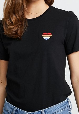 Woman wearing black t-shirt with small embroidered heart in red, beige, and blue stripes on left chest, paired with blue jeans.