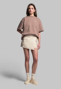 Brun oversized t-shirt med hvidt logo, parret med en beige omslagsnederdel med bånd og beige platform sneakers med teksturerede detaljer.