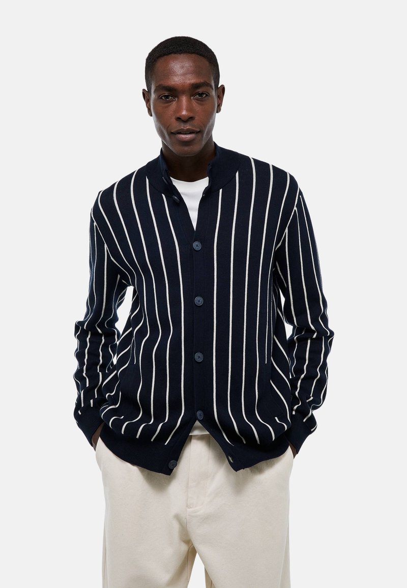Cardigan rayé marine en tissu tricot doux, avec des rayures verticales blanches, un col châle et une fermeture par boutons, associé à un pantalon clair.