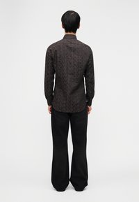 Άνδρας που στέκεται με την πλάτη στραμμένη, φοράει σκούρο πουκάμισο με μοτίβα και μαύρα τζιν με φαρδιά παντελόνια, μπροστά από απλό φόντο.