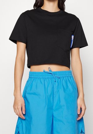 Camiseta negra corta con escote redondo, mangas cortas y un bolsillo en el pecho izquierdo. Presenta una etiqueta azul en el bolsillo. Combinada con pantalones cortos azules.