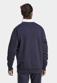 Mornar plavi dukserica s rebrastim manžetama i donjim rubom, s bijelim prugama na ramenima. Mekana tekstura, opušteni kroj i minimalistički dizajn.