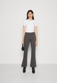 T-shirt bianco a maniche corte con logo rosso, abbinato a pantaloni svasati grigio scuro e stivaletti neri con tacco. Modella che tiene una piccola borsa nera.
