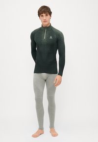 Camisola de performance de manga longa em verde escuro com fecho de quartzo, combinada com leggings texturizadas cinza claro com painéis nos joelhos.