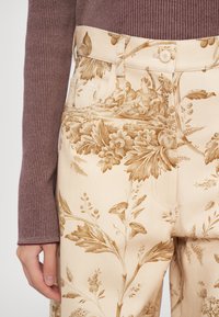 Pantaloni bej cu model floral, având o textură moale, cu ilustrații botanice maronii, talie înaltă și buzunare laterale. Se vede mâneca de pulover maro cu striații.