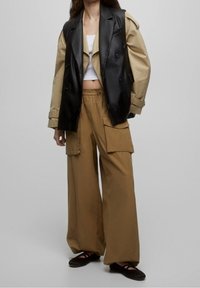 Gilet en cuir noir à double boutonnage porté sur une veste beige oversize, associé à un pantalon kaki  à jambes larges avec de grandes poches. Des ballerines noires complètent la tenue.