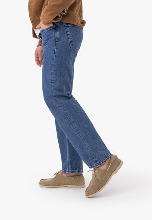 Persona con vaqueros azules, zapatos casuales de ante marrón claro y una chaqueta marrón, caminando con un pie ligeramente levantado.