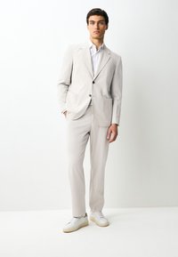 Junger Mann trägt einen hellbeigen Anzug mit weißem Hemd und weißen Turnschuhen, steht vor einem schlichten weißen Hintergrund.