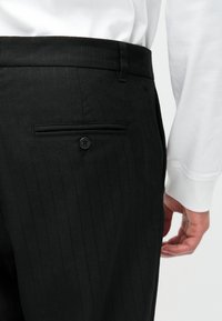 Fekete csíkos nadrág textúrális anyagból, hátul egy gombbal záródó zsebbel, fehér inggel párosítva.