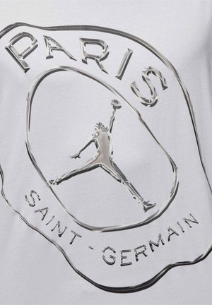 Logo argenté brillant du Paris Saint-Germain avec une silhouette stylisée en train de sauter, centré sur un fond en tissu blanc.
