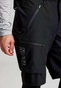 Pantaloni cargo neri con cerniere sulle tasche, realizzati con un tessuto testurizzato e un logo stampato in bianco sulla gamba sinistra.