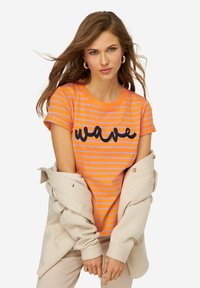Camiseta de rayas naranja y morada con un gráfico negro de "ola". Tiene mangas cortas y un escote redondo, combinada con una chaqueta beige.