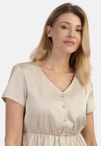 Beige lyhythihainen mekko V-päällyksellä, nappidetaililla ja laskeutuvalla vyötäröllä. Silkkinen kangas, jossa on hienovarainen kiilto.