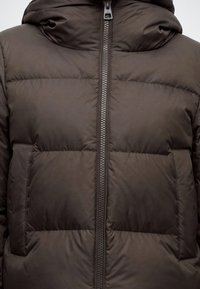 Gros plan d'une veste matelassée marron avec une fermeture éclair frontale et deux poches latérales, montrant le haut du torse et la région du cou.