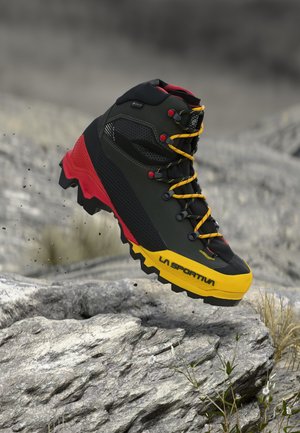 Schwarzer, roter und gelber La Sportiva-Wanderschuh mit robustem Profil, der über felsigem Gelände im Freien schwebt.