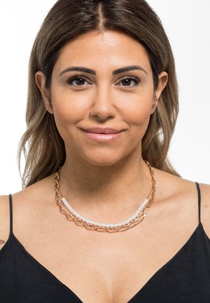 Gouden ketting met in elkaar verstrengelde schakels en meerdere rijen witte parels, gedragen op een blote sleutelbeen met een zwarte top.