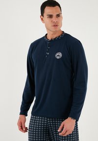 Námořnická dlouhá rukávová košile typu henley s knoflíkovou légou a kostkovaným límcem, v kombinaci s kostkovanými pyžamovými kalhotami v tmavých odstínech.