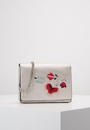 Kleine zilveren handtas met een kettingband en rood, roze en turquoise bloemborduursel aan de voorkant, tentoongesteld op een wit voetstuk.