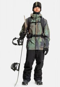 Snowboarder wearing a camo-print, waterproof jacket and black pants. Holds a snowboard and ski pole with black gloves and goggles.