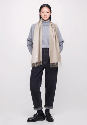 Beige Wollschal mit fransigen Enden, über einem grauen Rollkragenpullover getragen. Dunkelblaue Jeans mit aufgerollten Säumen und schwarzen Schuhen vervollständigen den Look.