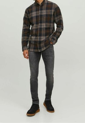 Homme portant une chemise à carreaux marron et grise à boutons, un jean slim gris foncé et des chaussures en daim noir avec des semelles beige, debout, une main dans la poche.