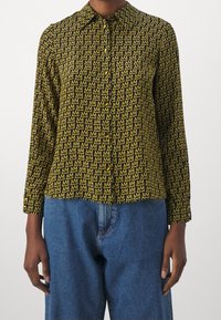 Camicia a righe nera e gialla con colletto, maniche lunghe e chiusura a bottoni, caratterizzata da motivi ripetuti a forma di 'M'. Abbinata a pantaloni in denim blu.