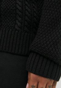 Hand met kruis tattoo's rustend nabij de zwarte gebreide sweatermanchet die textuurpatronen en ribbelranden toont.