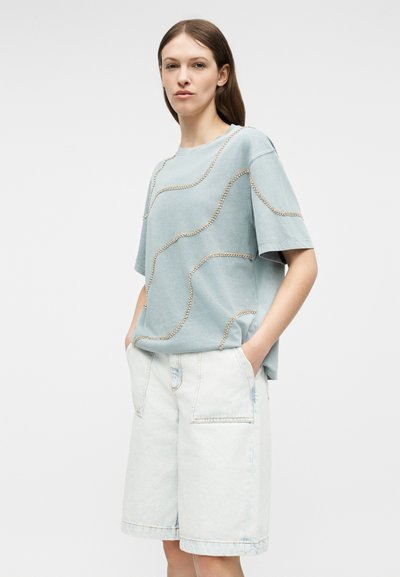 Γυναίκα που φοράει ανοιχτό μπλε φαρδιά κοντομάνικη μπλούζα με λεπτομέρειες από χάντρες και χαλαρά ανοιχτόχρωμα τζιν σορτς μέχρι το γόνατο με τσέπες.