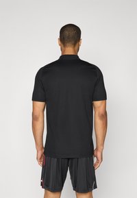 Sort kortærmet polo-shirt med krave, glat tekstur og en subtil glans. Bæres med sorte shorts med røde og hvide detaljer.