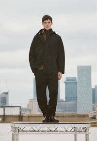 Sort quiltet jakke med høj krave, lagdelt over en brun skjorte. Sorte bukser og strømlinede sorte sko. Urbant baggrund, model på forhøjet platform.